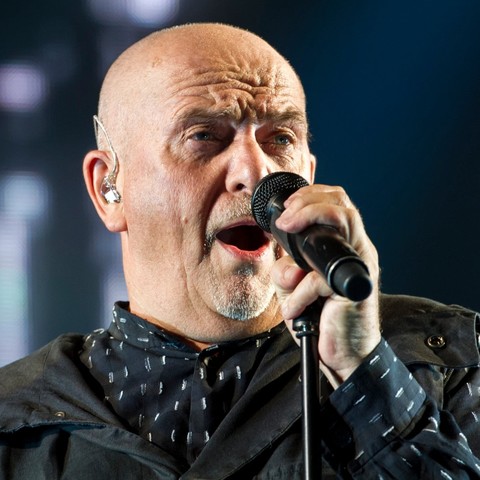Retrato de Peter Gabriel tomado desde la altura de sus hombros. Se observa con el micrófono en mano cerca de su boca mientras canta.