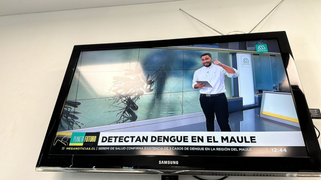Foto de la pantalla de un televisor con la imgen del noticiero donde se ve al presentador y una franja de texto en la parte inferior que dice "Detectan dengue en el Maule".