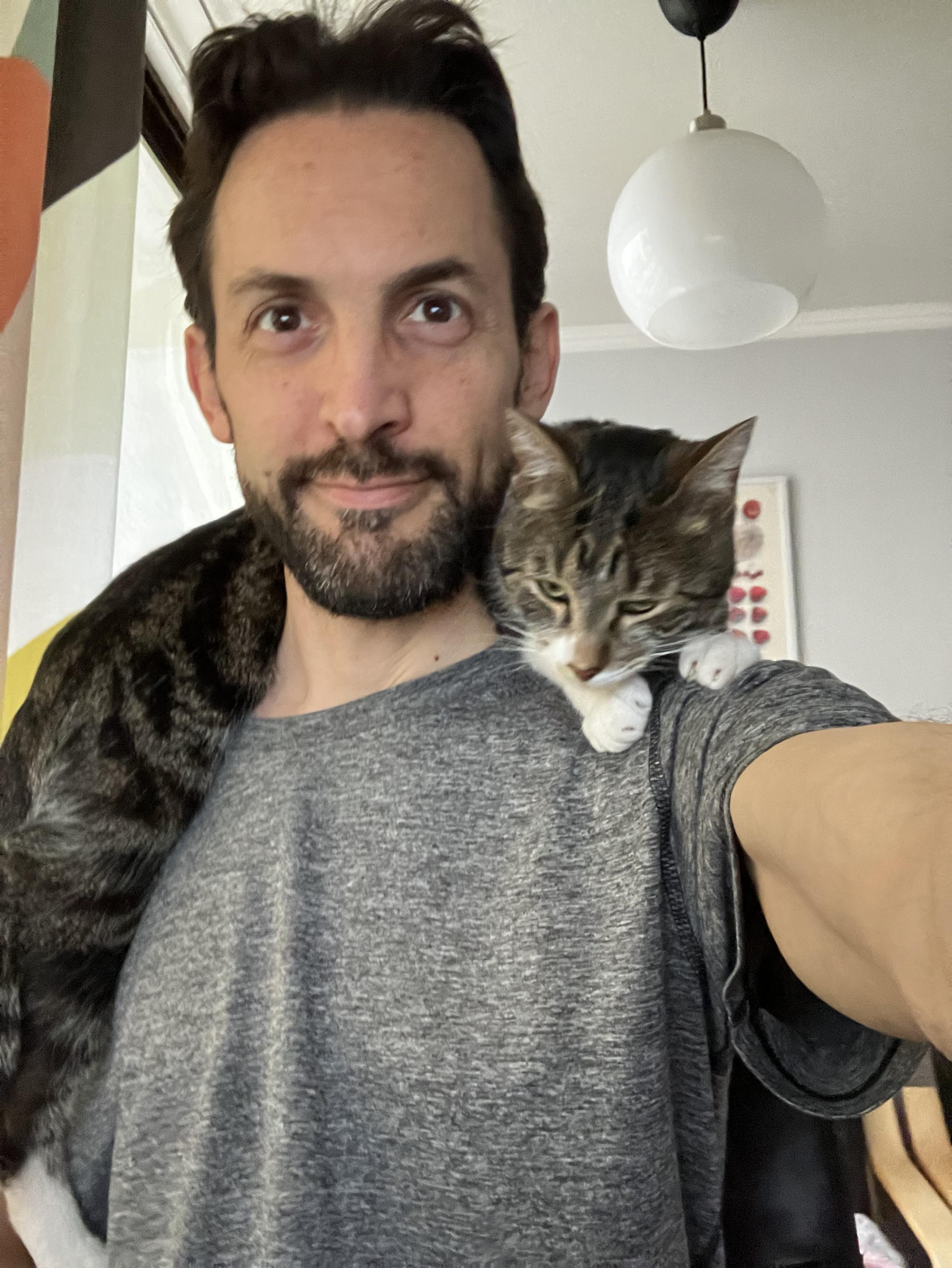 Sentado en mi escritorio, con una remera gris y un gato atigrado echado sobre mi cuello. Le encanta ponerse asรญ.