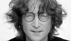 Retrato en primer plano en blanco y negro de John Lennon. Tiene el cabello oscuro, ondulado y de longitud media, algo despeinado, que cae sobre su frente y a los lados de su rostro. Lleva gafas redondas de montura fina. Mira directamente al frente con una expresión serena, mostrando una ligera sonrisa con los dientes superiores apenas visibles. Viste una prenda oscura, cuyo cuello es visible en la parte inferior de la imagen. El fondo es liso y claro.