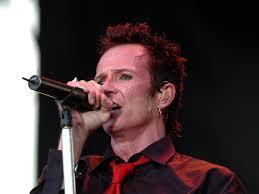 Scott Weiland pelo corto, rojizo y despeinado canta con una expresión intensa. Tiene los ojos azules y la boca abierta como si estuviera gritando o cantando una nota alta, con la frente ligeramente arrugada. Lleva una camisa oscura, que parece negra o burdeos, y una corbata roja brillante. Sostiene un micrófono negro con la mano derecha, apoyado en un soporte plateado. Un pequeño pendiente de aro cuelga de su oreja izquierda. El fondo es oscuro y difuminado, sugiriendo un escenario de concierto.