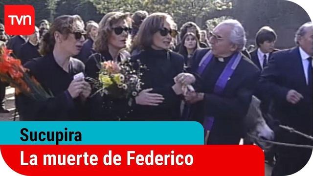 Una captura de pantalla de un episodio de televisión de TVN, titulado "Sucupira: La muerte de Federico". La imagen muestra una escena de funeral al aire libre durante el día. En primer plano, varias mujeres vestidas de luto, con ropa oscura y gafas de sol, están reunidas. La mujer más a la izquierda lleva un gran ramo de flores rojas y naranjas y parece angustiada. En el centro, una mujer con un ramo de flores más claras se encuentra junto a otra mujer con un cuello alto negro, quien tiene sus …