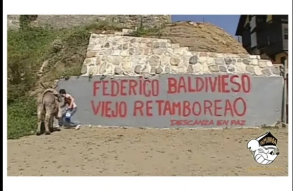 Una imagen al aire libre muestra un burro y una persona junto a un muro de contención. El muro, de color gris claro, tiene un graffiti grande en letras rojas que dice "FEDERICO BALDIVIESO VIEJO RETAMBOREAO". Debajo, en letras más pequeñas, se lee "DESCANZA EN PAZ". Sobre este muro hay otra sección de pared construida con piedras irregulares y vegetación. A la izquierda, un burro de color marrón claro se encuentra de espaldas a la cámara. Junto a su parte trasera, una persona se agacha o arrodil…