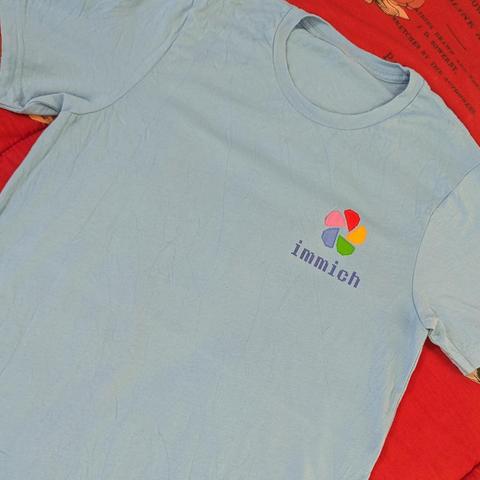 Una camiseta de color azul claro, ligeramente arrugada y extendida sobre una superficie. El fondo es una tela roja con texto impreso, posiblemente páginas de un libro antiguo o un patrón. En la parte superior izquierda del pecho de la camiseta (vista desde el frente), hay un logotipo bordado. Consiste en un símbolo multicolor que parece una flor estilizada o un diafragma de cámara, formado por seis segmentos en forma de pétalos o aspas, cada uno de un color vibrante: rojo, naranja, amarillo, ve…