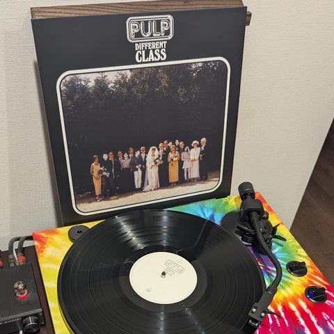 La imagen muestra un tocadiscos con un disco de vinilo negro reproduciéndose, y la portada de un álbum de vinilo apoyada detrás. La portada, de color negro, lleva el título "PULP DIFFERENT CLASS" en la parte superior. En el centro, hay una fotografía enmarcada de un grupo de personas vestidas de forma formal, incluyendo una novia de blanco y un novio, posando al aire libre frente a árboles oscuros, simulando una foto de boda. El tocadiscos tiene una base de colores vivos estilo tie-dye, con ton…