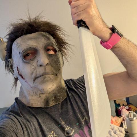 Una selfie de cerca de una persona que lleva una máscara de Michael Myers. La máscara es de color gris pálido y tiene un aspecto sucio o envejecido, con cabello oscuro y despeinado en la parte superior. Hay una herida falsa rojiza visible en la mejilla derecha de la máscara. La persona mira directamente a la cámara y sostiene un cuchillo grande de utilería, de color blanco, con la mano derecha levantada. En su muñeca derecha lleva un reloj inteligente de color rosa brillante. Viste una camiseta…
