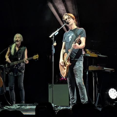 Imagen de un concierto en vivo con dos músicos en el escenario. En primer plano a la derecha, un hombre con gafas y pelo castaño claro canta en un micrófono mientras toca una guitarra eléctrica de color madera clara. Viste una camiseta oscura con un gráfico claro y jeans. A su izquierda y ligeramente detrás, otro hombre con pelo largo y rubio o blanco toca un bajo eléctrico negro y también canta en un micrófono. Viste un chaleco oscuro sin mangas y pantalones oscuros. El fondo es oscuro, ilumin…