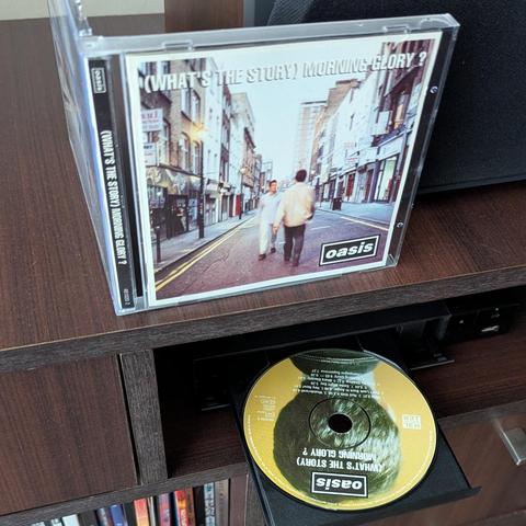 Una imagen muestra el icónico álbum de Oasis, "(WHAT'S THE STORY) MORNING GLORY?", en dos formatos. En la parte superior, el estuche del CD está de pie sobre un mueble de madera oscura. La portada del álbum muestra a dos hombres caminando de espaldas por el centro de una calle urbana con edificios a ambos lados y el título en la parte superior. En la parte inferior derecha, el disco dorado del mismo álbum, con el título y el logo de Oasis visibles, está parcialmente fuera de la bandeja de un re…