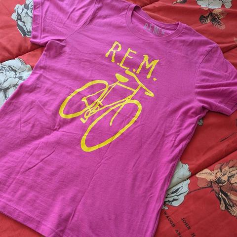 Una camiseta de color fucsia intenso, extendida sobre una superficie con un patrón floral. La camiseta lleva el logotipo "R.E.M." en letras amarillas brillantes en la parte superior, y debajo, el contorno de una bicicleta en amarillo brillante. La superficie de fondo es de color rojo con grandes diseños florales en tonos blancos, grises y naranjas.