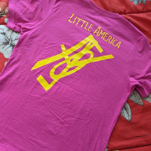Una camiseta de manga corta de color rosa fucsia brillante, extendida sobre una superficie con un patrón floral rojo y blanco. En la parte superior trasera de la camiseta, cerca del cuello, se lee la frase "LITTLE AMERICA" en letras mayúsculas de color amarillo. Debajo de la frase, hay un gran gráfico abstracto de color amarillo que parece consistir en letras estilizadas y entrelazadas.