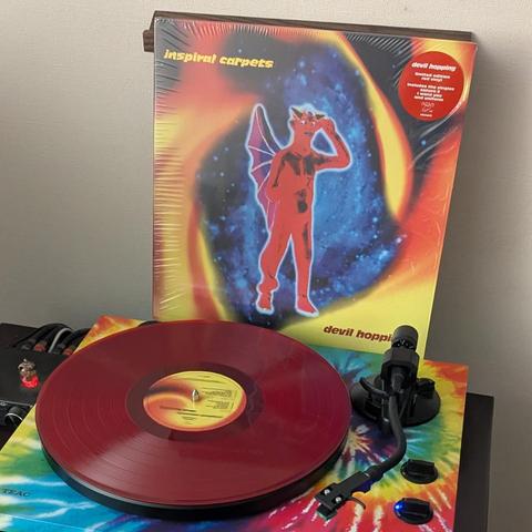 La imagen muestra una escena musical con un disco de vinilo y su portada. La portada del álbum "devil hopping" de Inspiral Carpets se apoya verticalmente. Presenta una figura roja alada y con cuernos, similar a un diablo, de pie con una mano en la cabeza. El fondo es un vibrante patrón cósmico o nebulosa en tonos azules, púrpuras, naranjas y amarillos. Un sticker rojo en la esquina superior derecha de la portada anuncia que es una edición limitada en vinilo rojo.

Delante de la portada, sobre u…