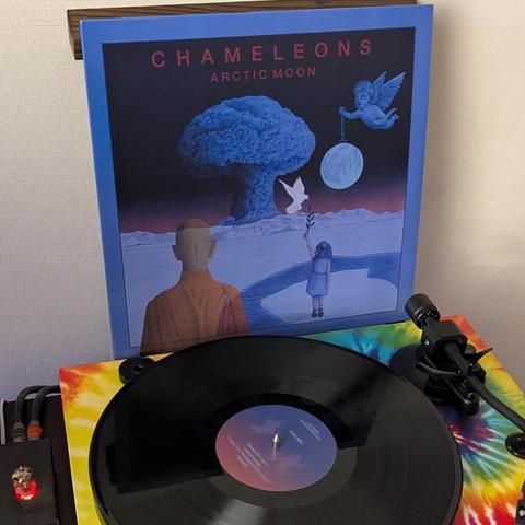 La imagen muestra un tocadiscos con un vinilo negro en primer plano, y la portada de un álbum de música detrás. La portada del álbum, titulada "CHAMELEONS - ARCTIC MOON", presenta una escena surrealista y onírica dominada por tonos azules y morados oscuros. En el centro, hay una gran nube azul en forma de hongo. A la izquierda, un hombre calvo con una túnica naranja le da la espalda al espectador, mirando la nube. A la derecha, una niña con un vestido azul claro sostiene una rama con hojas, mie…