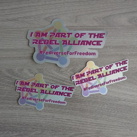 Tres pegatinas idénticas se muestran sobre una superficie de madera de tono claro. Cada pegatina presenta un diseño irregular con una red de puntos interconectados en tonos pastel (naranja, amarillo, verde, azul, morado) como fondo. Sobre este fondo, el texto principal en mayúsculas de color magenta o morado intenso dice: 'I AM PART OF THE REBEL ALLIANCE'. Debajo de este, en el mismo color, aparece el hashtag: '#FediverseForFreedom'.