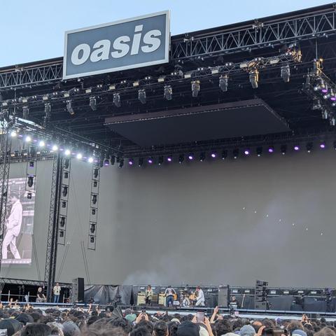 Una vista frontal de un gran escenario de concierto al aire libre bajo un cielo azul claro. En la parte superior del escenario, un gran cartel rectangular blanco y gris muestra el nombre "Oasis" en letras mayúsculas. El escenario cuenta con una compleja estructura de andamios metálicos (trussing) de la que cuelgan numerosas luces, algunas emitiendo un brillo púrpura, y varias pantallas. Una gran pantalla vertical a la izquierda muestra a una persona en ropa clara, mientras que la pantalla princ…
