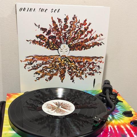 Una imagen muestra un disco de vinilo negro girando sobre un tocadiscos con un llamativo patrón tie-dye de colores rojo, naranja, amarillo, verde y azul. Detrás del tocadiscos, se alza la portada del álbum. La portada es blanca y presenta una ilustración central de un rostro estilizado y pálido con los ojos cerrados y marcas doradas en la frente. De este rostro emanan grandes masas de formas onduladas y fluidas en tonos marrones, naranjas y dorados, que se extienden tanto hacia arriba como haci…