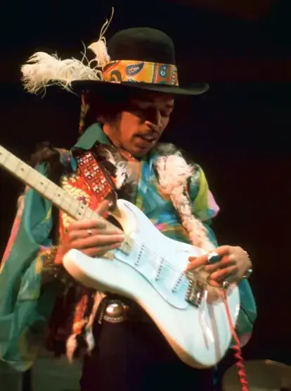 Jimi Hendrix en un escenario oscuro, absorto tocando su guitarra eléctrica. Lleva un sombrero de ala ancha negro adornado con una banda de patrón paisley en tonos vibrantes (naranjas, amarillos, azules) y plumas blancas. Su camisa es multicolor, principalmente turquesa y azul, complementada con un chaleco o adorno peludo en blanco y negro sobre los hombros. La correa de su guitarra es roja con detalles geométricos. En sus manos, se aprecian varios anillos. La guitarra es una Fender Stratocaster…