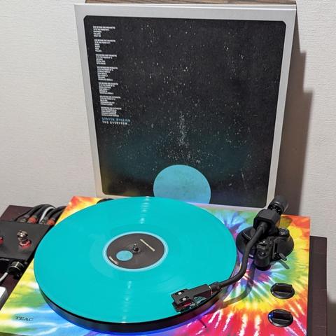 Un disco de vinilo de color turquesa brillante gira en un tocadiscos TEAC. El tocadiscos tiene un vibrante tapete con estampado tie-dye, mostrando un remolino de colores como rojo, amarillo y azul. Detrás, se apoya la carátula de un álbum con un fondo oscuro que simula el espacio estrellado y un planeta azul claro en la parte inferior. En la carátula se ve texto blanco, incluyendo el nombre del artista "Steven Wilson" y una lista de canciones.