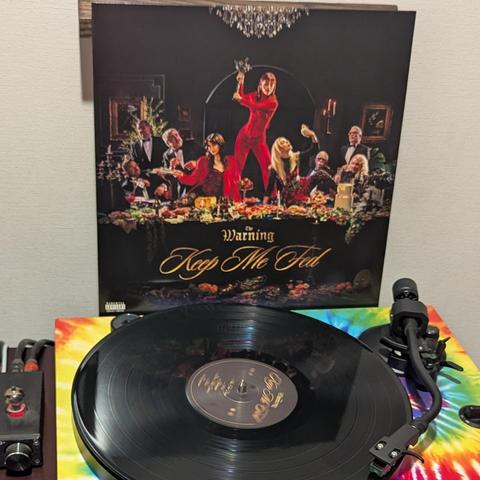 Una imagen muestra un disco de vinilo negro girando en un tocadiscos, con su portada apoyada detrás.

La portada, de fondo oscuro, presenta una opulenta cena de gala. Una mujer vestida con un mono rojo brillante se alza de pie sobre la mesa, levantando un ramo de flores como si fuera un arma. Alrededor de la mesa, ricamente decorada con comida, bebida y candelabros, se sientan otras dos mujeres y varios hombres, todos con atuendos elegantes. Las dos mujeres sentadas también visten tonos rojos y…