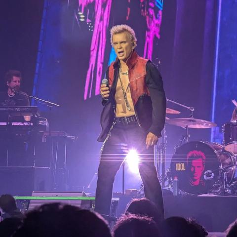 Una imagen de Billy Idol actuando en un concierto en vivo. El cantante está en el centro del escenario, con su característico pelo rubio y puntiagudo. Viste una chaqueta abierta de cuero rojo y negro, sin camisa, mostrando su torso. Lleva un collar largo de cadena plateada con un colgante. Sostiene un micrófono con su mano derecha y tiene la boca abierta, cantando con intensidad. A su izquierda, un tecladista con barba está sentado tocando un sintetizador. A su derecha, se ve parte de una bater…
