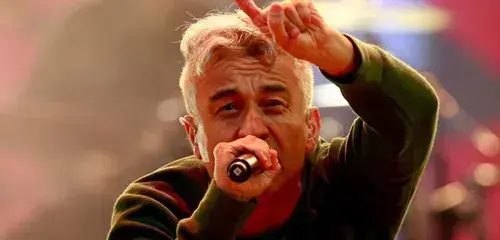 Jorge González cabello canoso y peinado hacia arriba canta con expresión intensa en un escenario. Sostiene un micrófono con una mano mientras levanta la otra haciendo el gesto de cuernos de rock. Viste un suéter verde oscuro. El fondo es borroso con luces cálidas y un ambiente brumoso.
