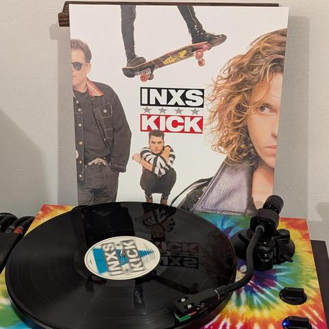 Una imagen muestra un disco de vinilo del álbum "Kick" de INXS en un tocadiscos, con la carátula del álbum en la parte trasera. La carátula, predominantemente blanca, presenta a miembros de la banda en poses estilizadas: unas piernas en patineta en la parte superior, un hombre con gafas de sol y chaqueta vaquera a la izquierda, otro en cuclillas con una camiseta a rayas en el centro inferior, y un primer plano del rostro de Michael Hutchence con pelo rizado y chaqueta de cuero a la derecha. El …