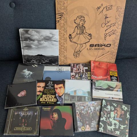 Una colección de álbumes de música y un libro sobre una superficie de tela oscura, posiblemente un sofá o alfombra.

En la parte superior derecha, hay una caja grande de cartón color café claro, con la ilustración de una niña caricatura saltando la cuerda. En ella se lee "SAIKO LAS HORAS" y varias firmas manuscritas.

A su izquierda, un libro de tapa dura en blanco y negro, mostrando un paisaje con un cielo nublado, una montaña a lo lejos y un terreno texturizado.

Debajo de estos, se apilan nu…