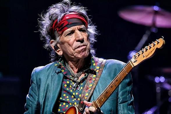 Primer plano de Keith Richards, guitarrista de The Rolling Stones, tocando una guitarra eléctrica en un escenario oscuro. Tiene el pelo gris y revuelto, lleva una bandana roja estampada y una chaqueta verde azulado sobre una camisa oscura con puntos de colores. Su rostro arrugado muestra una expresión concentrada, con los labios ligeramente fruncidos. Cuelgan varias cadenas de su cuello. Sostiene la guitarra con una correa ancha y colorida, y parte del mástil y la pala de la guitarra son visibl…