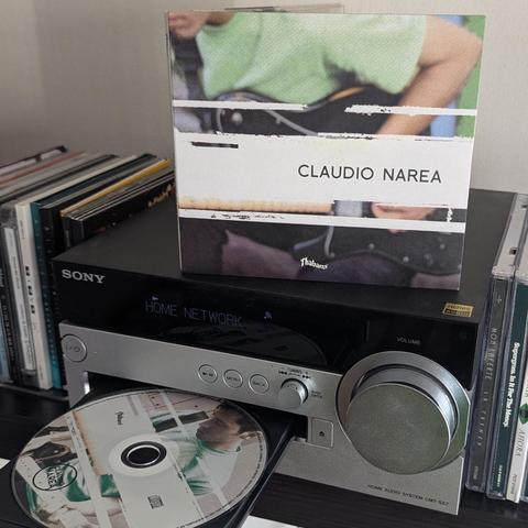Una imagen muestra un sistema de audio doméstico Sony de color negro y plateado. Sobre la unidad principal negra, hay una carátula de CD en posición vertical. La carátula es del artista "CLAUDIO NAREA" y muestra una imagen de una persona con una camisa verde tocando una guitarra, con un efecto de bandas horizontales blancas que atraviesan la foto. El reproductor de CD, que tiene una bandeja plateada abierta, está a mitad de camino insertando un disco. El disco también tiene la carátula de "CLAU…
