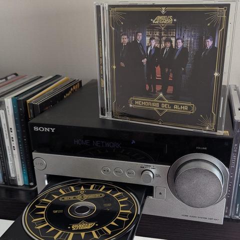 Una imagen de un sistema de audio doméstico Sony CMT-SX7 de color negro y plateado. El reproductor de CD tiene la bandeja abierta, mostrando un CD negro con detalles dorados de 'Los Ángeles Negros - Memorias del Alma' parcialmente insertado. Encima del sistema de audio, está la caja del mismo álbum, de pie. La carátula del CD muestra a seis hombres vestidos con trajes oscuros, de pie frente a una reja decorativa, con un borde dorado estilo art déco y el título 'Memorias del Alma'. A la izquierd…