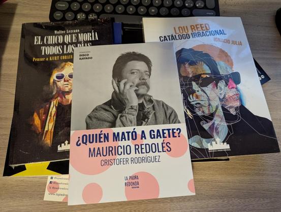 La imagen muestra tres libros apilados sobre una superficie de madera clara, con un teclado de computadora oscuro desenfocado al fondo.

El libro de la izquierda, de tapa oscura, se titula "EL CHICO QUE MORÍA TODOS LOS DÍAS: Pensar a KURT COBAIN" de Walter Lezcano. Muestra una ilustración de Kurt Cobain con gafas de sol blancas y cabello largo.

En el centro, el libro blanco se titula "¿QUIÉN MATÓ A GAETE?" de Mauricio Redolés y Cristofer Rodríguez, con una foto en blanco y negro de un hombre c…