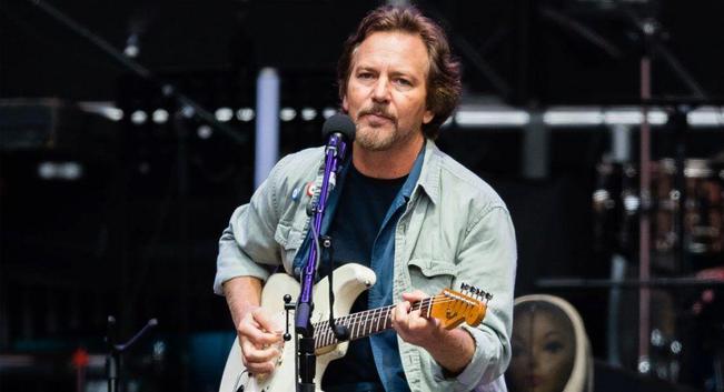 El músico Eddie Vedder, un hombre caucásico de mediana edad con cabello castaño ondulado y barba, está de pie en el escenario tocando una guitarra eléctrica de color crema. Viste una camisa de trabajo de color verde claro sobre una camiseta oscura y sostiene una púa para tocar las cuerdas. Canta en un micrófono negro montado en un soporte violeta. El fondo es oscuro y ligeramente desenfocado, sugiriendo un entorno de concierto con luces y equipo de escenario apenas visibles.