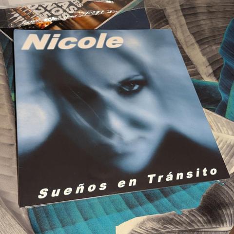 La portada de un álbum musical muestra un primer plano estilizado de un rostro femenino con un efecto borroso y etéreo, dominado por tonos azules grisáceos y negros. Se distingue claramente un ojo (el derecho de la persona) con una mirada intensa, mientras el resto del rostro se difumina en la oscuridad. En la parte superior, se lee el nombre "Nicole" en letras blancas y grandes. En la parte inferior, el título "Sueños en Tránsito" también en letras blancas. El álbum descansa sobre una superfic…