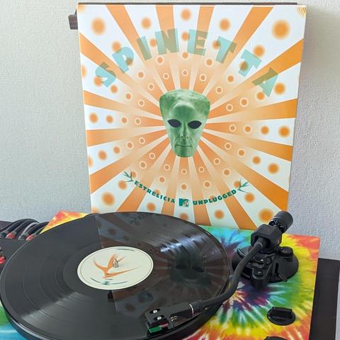 Una imagen de un disco de vinilo en un tocadiscos, con la carátula del álbum detrás. La carátula es blanca con rayos naranjas y círculos que irradian desde el centro, donde se encuentra una cabeza de alienígena verde con ojos grandes y negros. Arriba, el título "SPINETTA" en letras celestes, y abajo, "ESTRELICIA" y "UNPLUGGED" en verde, separados por el logo de MTV. En primer plano, un tocadiscos negro reproduce un disco de vinilo negro. El centro del disco tiene una etiqueta blanca con un dise…