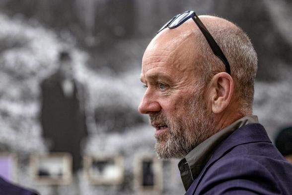 Michael Stipe con barba canosa y cabeza calva se muestra de perfil, mirando hacia la derecha de la imagen. Lleva gafas de montura oscura sobre la parte superior de su cabeza. Viste una chaqueta de traje azul oscuro a rayas y una camisa más oscura. El fondo está desenfocado, revelando una gran fotografía en blanco y negro de una figura humana (posiblemente un hombre de traje) y algunas imágenes más pequeñas enmarcadas debajo, lo que sugiere un entorno de galería o exposición.