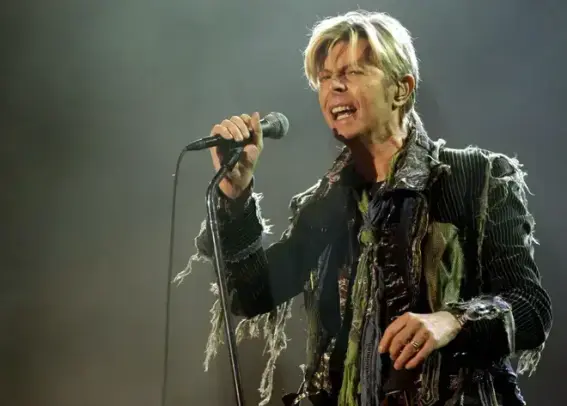 David Bowie canta apasionadamente en un escenario. Tiene el cabello rubio claro, peinado hacia atrás, y una expresión enérgica con la boca abierta y los ojos ligeramente entrecerrados. Viste una chaqueta oscura a rayas con un diseño deshilachado y artístico, con numerosos flecos y rasgaduras. Debajo, se aprecian capas de tela en tonos oscuros y verdosos. Sostiene un micrófono con su mano izquierda. El fondo es oscuro y difuso, iluminado tenuemente, lo que sugiere un ambiente de concierto.