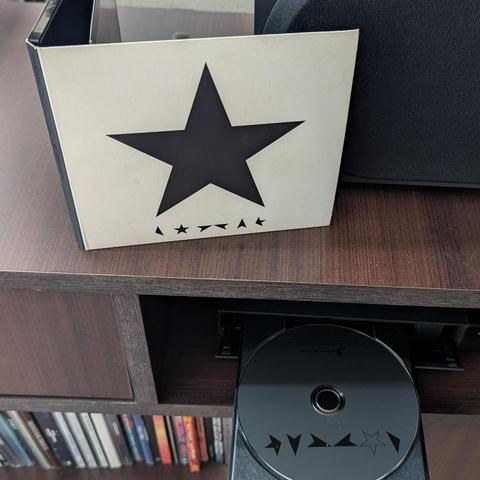 Una portada de álbum de color crema con una gran estrella negra de cinco puntas en el centro, y una fila de pequeños símbolos negros (estrellas y formas de flecha) debajo, está de pie sobre una superficie de madera oscura. Debajo, un CD negro con un patrón de símbolos similar (incluyendo una estrella delineada) emerge parcialmente de un reproductor de discos. La imagen muestra una estantería de madera oscura, con una sección inferior llena de lomos de otros CDs o libros, y parte de un altavoz n…