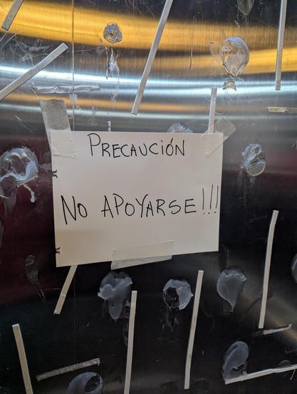 Un cartel blanco escrito a mano está pegado con cinta adhesiva a una superficie vertical, que parece ser metálica y muy desgastada. El cartel dice "PRECAUCIÓN" en la parte superior y "NO APOYARSE!!!" en la inferior, ambos escritos en mayúsculas con rotulador negro. La superficie de fondo está muy rayada y cubierta de numerosos restos de pegamento seco y pequeñas tiras de material claro adheridas, con la parte superior de un tono plateado o dorado cepillado y la inferior de un color más oscuro, …