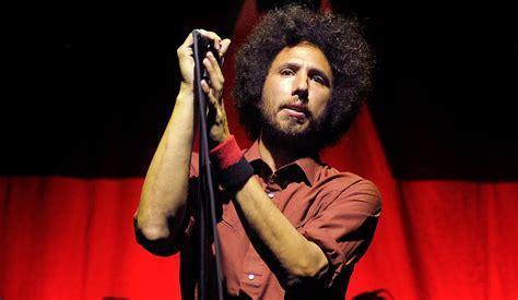 Zack de la Rocha con cabello afro oscuro y rizado, y barba, sostiene un micrófono con ambas manos en un escenario. Viste una camisa de botones color óxido y una muñequera roja y negra en su muñeca izquierda. Su mirada se dirige ligeramente hacia arriba y al frente, mientras el fondo es oscuro, iluminado con tonos rojos dramáticos que sugieren un concierto o actuación en vivo.