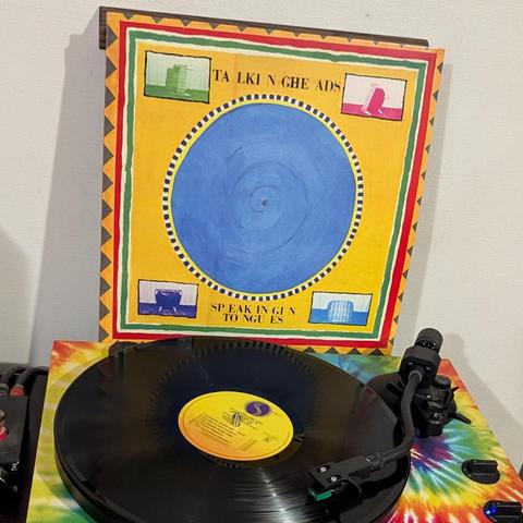 Una imagen de un tocadiscos reproduciendo un disco de vinilo, con la carátula del álbum 'Speaking in Tongues' de Talking Heads detrás. La carátula tiene un fondo amarillo brillante, enmarcado por un borde grueso con patrones geométricos en rojo, verde, amarillo y negro. En el centro hay un gran círculo azul, rodeado por una línea punteada negra, y cuatro pequeños recuadros con ilustraciones abstractas en tonos verdes, rosas y azules. Debajo, el disco de vinilo negro gira en un tocadiscos cuya b…