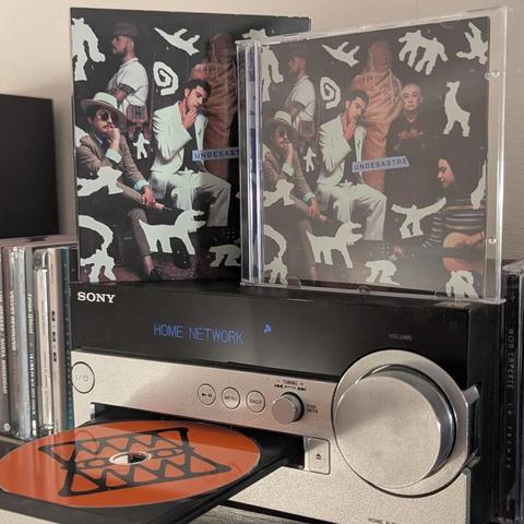 Una vista en primer plano de un sistema de audio doméstico Sony con varios CDs. En la parte superior, hay dos copias del álbum "UNDESASTRE" con una portada idéntica. La portada es oscura y presenta a varias personas, incluyendo dos hombres en primer plano (uno con sombrero y otro con traje claro), rodeados de figuras abstractas blancas que parecen animales y símbolos. La versión en caja de plástico transparente de CD también muestra a una mujer tocando la guitarra a la derecha.

Debajo de los á…