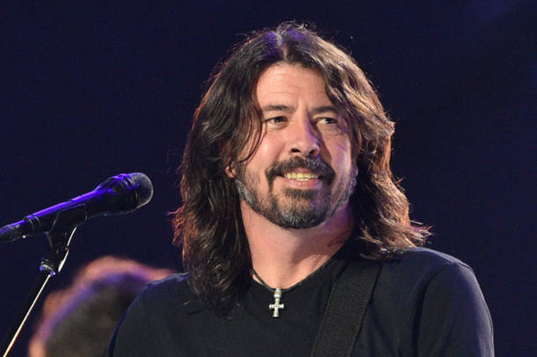 Una imagen de cerca de Dave Grohl sonriendo ampliamente, mirando ligeramente hacia su derecha. Tiene el pelo largo castaño oscuro y ondulado que le cae sobre los hombros, y una barba y bigote entrecanos. Viste una camiseta negra y lleva un collar con un colgante de cruz plateada sobre su pecho. Una correa de guitarra negra es visible sobre su hombro derecho. A su izquierda, se aprecia la parte superior de un micrófono oscuro. El fondo es borroso y oscuro, con iluminación tenue en tonos morados …