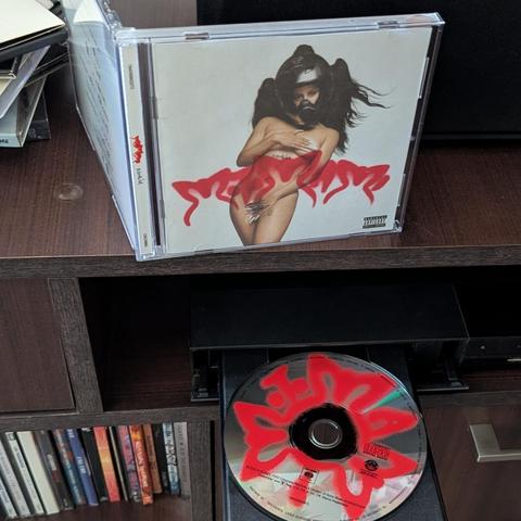 Una caja de CD del álbum 'Motomami' de Rosalía está de pie sobre una estantería de madera oscura. La portada muestra a una figura femenina semidesnuda con un casco de motocicleta oscuro y largo cabello, con sus manos cubriendo estratégicamente sus pechos y el área pélvica. Su cuerpo está adornado con formas rojas estilizadas que parecen llamas o alas. En la parte inferior de la imagen, un disco plateado del álbum 'Motomami' se encuentra en la bandeja abierta de un reproductor de CD. El disco ti…