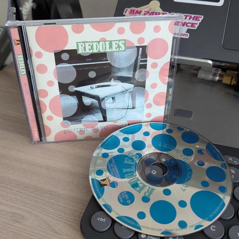 La imagen muestra el álbum de CD de Mauricio Redoles, titulado "¿Quién mató a Gaete?", con la caja y el disco fuera de ella, sobre un escritorio de madera clara.

La carátula del CD tiene un fondo crema decorado con numerosos círculos o puntos rosas de distintos tamaños. En el centro, hay una fotografía en blanco y negro de una mesa pequeña y una silla antigua, con un objeto conectado por un cable sobre la mesa. El nombre del arista "REDOLES", aparece en letras grandes de color verde en la part…