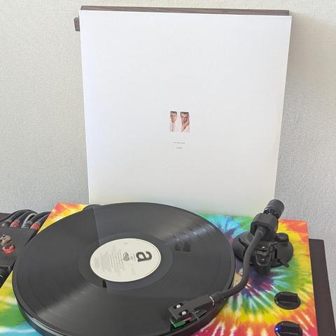 Un tocadiscos con un disco de vinilo negro sobre una base de colores vibrantes con estampado tie-dye (rojo, amarillo, verde, azul). Detrás, se alza la carátula de un álbum blanca, que muestra dos pequeñas fotografías idénticas del rostro de un hombre. En el centro del disco, hay una etiqueta blanca con la letra 'a' en negro, y el brazo de lectura negro con un detalle verde está sobre el disco. El fondo es una pared lisa de color claro."