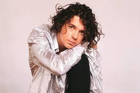 Michael Hutchence con cabello oscuro, abundante y rizado, mira directamente a la cámara. Viste una camisa de manga larga plateada brillante y pantalones oscuros. Está en una pose relajada, apoyando su mejilla en su brazo doblado o rodilla, con una expresión seria o pensativa. El fondo es liso y de color claro, casi blanco o beige rosado.