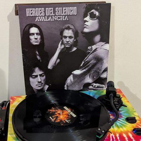 La imagen muestra un disco de vinilo negro del álbum "Avalancha" de Héroes del Silencio, colocado sobre un tocadiscos. Detrás, se aprecia la portada del álbum, que presenta una fotografía en blanco y negro de los cuatro miembros de la banda con expresiones serias. Arriba de la portada, se lee claramente "HÉROES DEL SILENCIO" y "AVALANCHA". El tocadiscos destaca por su vibrante base con un diseño de espiral tie-dye en tonos arcoíris, incluyendo rojo, naranja, amarillo, verde y azul. El vinilo es…