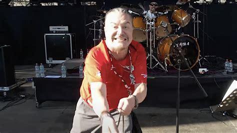 Mike Patton con el pelo recogido en una coleta y barba, sonríe ampliamente con una expresión muy enérgica y alegre, mirando directamente a la cámara. Viste una camiseta roja y pantalones grises, y se inclina hacia adelante con los brazos extendidos. Está en un escenario, con un telón de fondo oscuro. Detrás de él, a la derecha, se ve a un baterista con gafas de sol sentado frente a una batería de color madera con el logo "TAMA". En el escenario también se aprecian varias botellas de agua y equi…