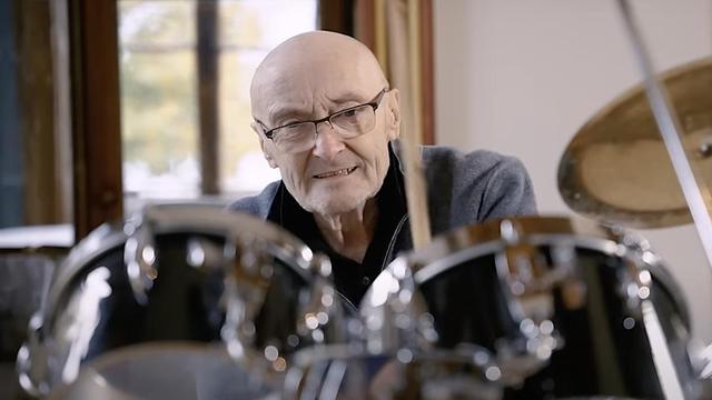 Phil Collins calvo y con gafas de montura oscura, aparece en primer plano tocando la batería. Su rostro, en el centro de la imagen y con una expresión concentrada o de esfuerzo, mira hacia los tambores. Viste una chaqueta gris sobre una camisa oscura y sostiene una baqueta sobre uno de los tambores negros y brillantes con herrajes cromados. A la derecha, se vislumbra un platillo dorado. El fondo es un interior desenfocado con una ventana a la izquierda.