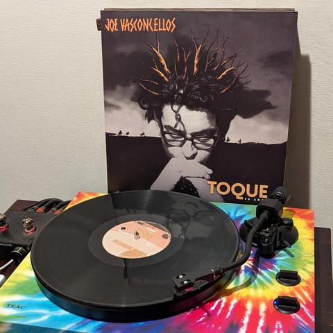 Un tocadiscos con un disco de vinilo negro se muestra en primer plano, con la carátula del álbum "Toque" de Joe Vasconcellos apoyada detrás. La carátula presenta un retrato en blanco y negro del artista, un hombre con gafas que mira pensativamente hacia abajo con la barbilla apoyada en su mano. Su cabello oscuro está peinado con mechones puntiagudos de color naranja-marrón que se extienden hacia arriba. La base del tocadiscos tiene un vibrante diseño tie-dye con colores como rojo, naranja, amar…