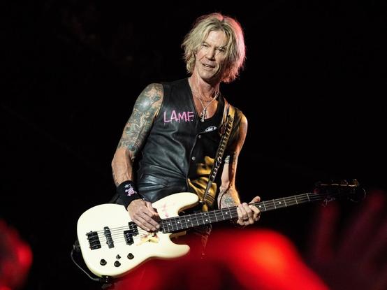 Duff Mckagan, un hombre rubio de pelo largo, con un chaleco de cuero negro sin mangas y brazos tatuados, sonríe levemente mientras toca un bajo eléctrico de color blanco crema. El bajo tiene un golpeador negro y un pequeño adhesivo amarillo cerca del puente. El hombre está en una pose de actuación contra un fondo oscuro, con una iluminación roja tenue visible en la parte inferior derecha, sugiriendo un escenario de concierto.